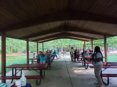 People gathered under a pavilion with picnic tables in a park.