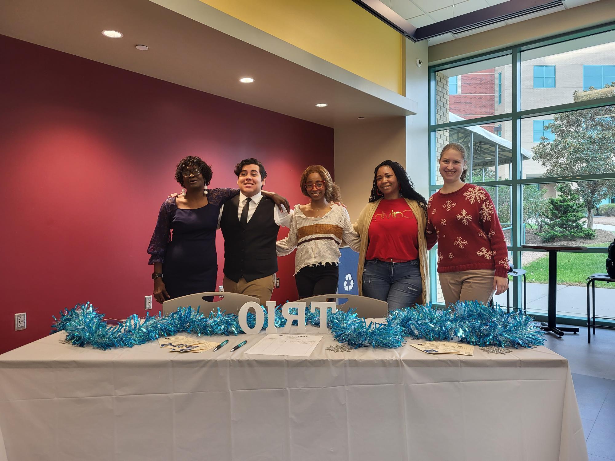 A group of five people stand in a room behind a table with "TRIO" letters and blue tinsel.